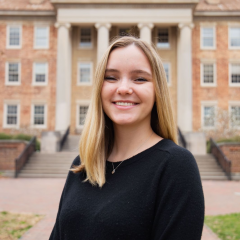 Picture of Graduate Research Assistant Devin Wilson. She has blond hair, is wearing a black sweater, and is standing outside in front of a building.